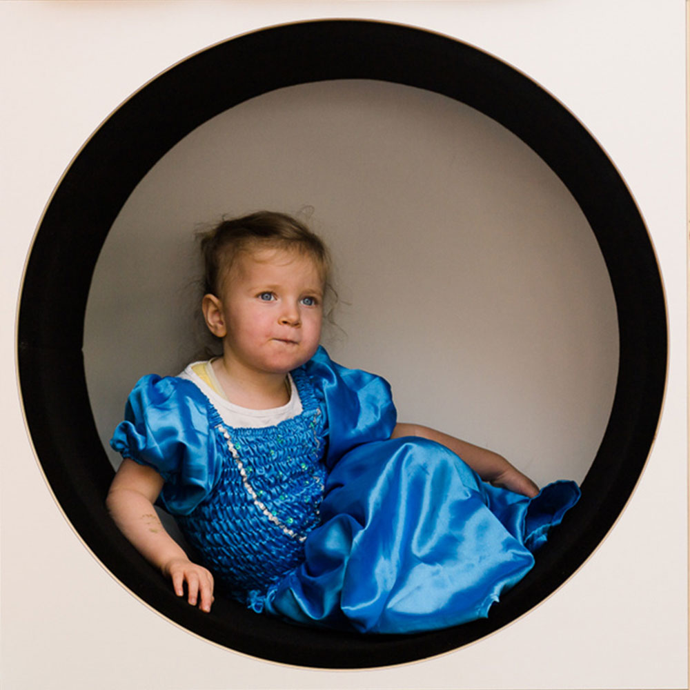 Little girl in a blue dress sitting in a round cubby hole at Spotted Frog Preschool