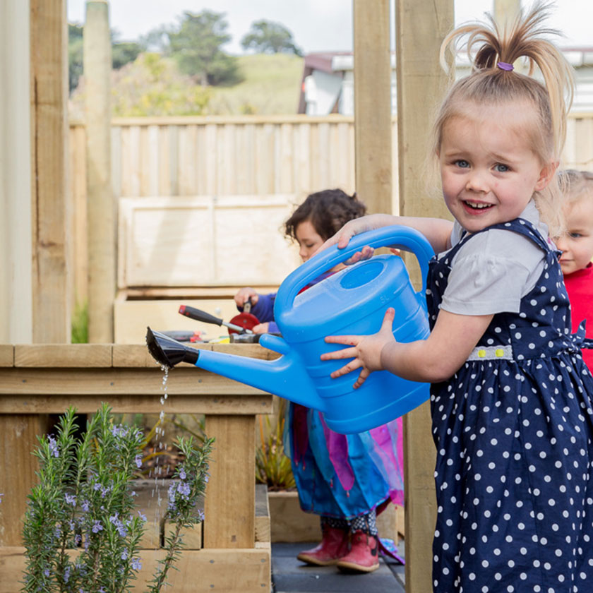 A preschool girl watering plants with a blue watering can at Spotted Frog Preschool